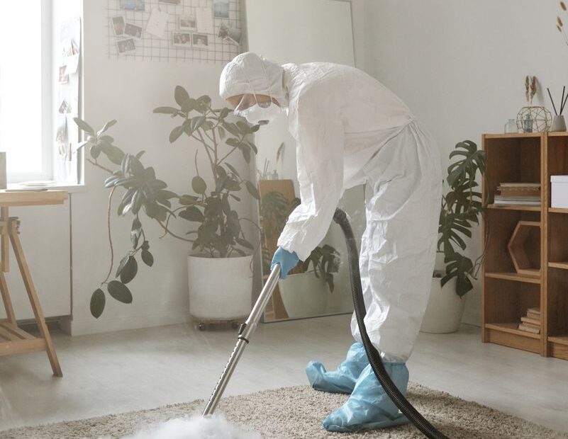 A person in full protective gear disinfects a carpet in a bright indoor space.